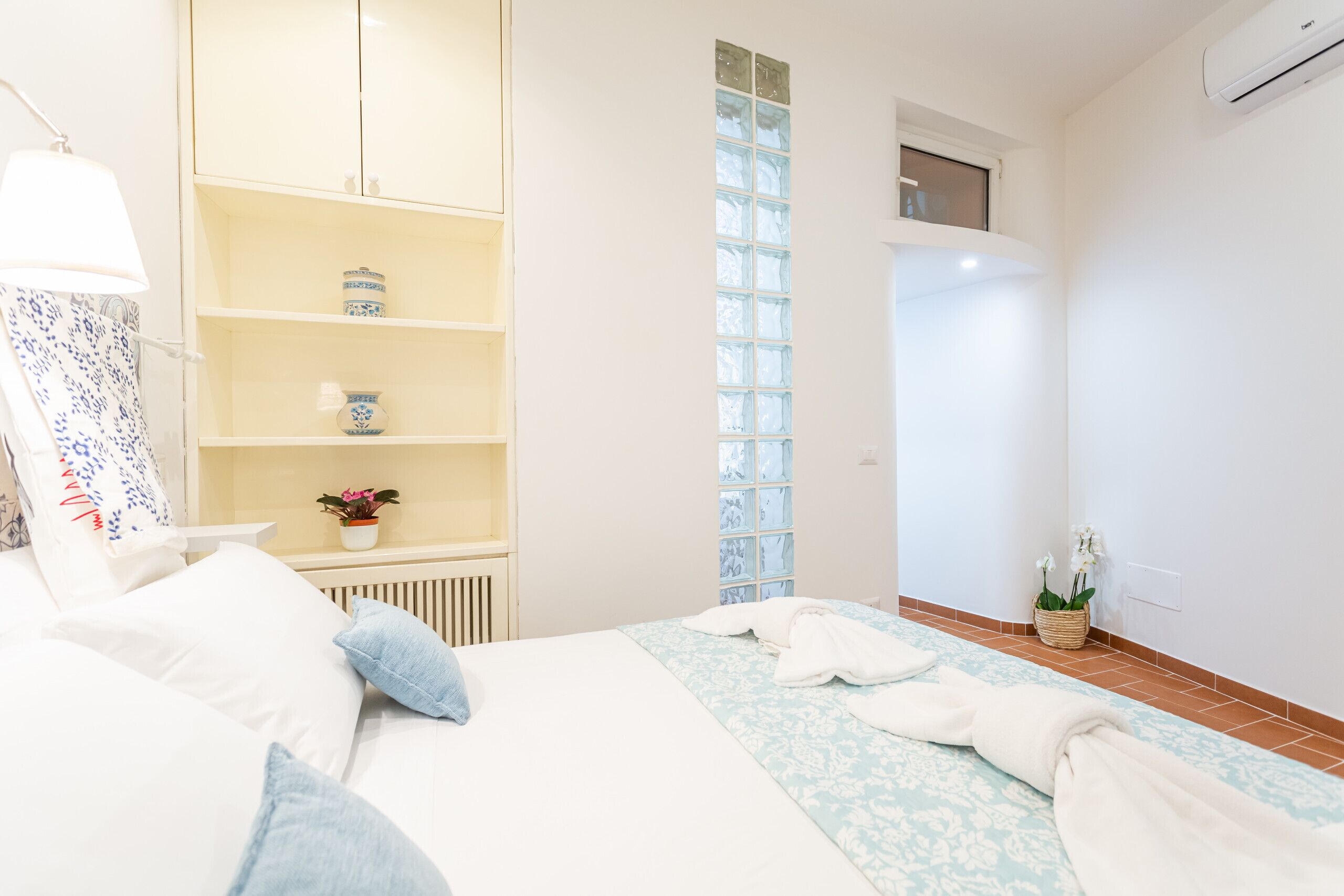 A hotel room with a bed, a bedside table, a lamp, a shelf with decorative items, a window, and a potted plant.