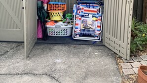 A storage shed with a metal door, containing various items like baskets, a stroller, and a stroller cover.