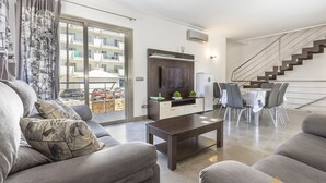 A modern living room with a sofa, coffee table, and a TV on a wall. There's a dining area with a table and chairs. A staircase is visible in the background. - Villa Albufera en Playa Muro by Mallorca North Villas (Platja de Muro)