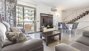 Uma sala de estar moderna com sofá, mesa de centro e televisão na parede. Há uma área de jantar com mesa e cadeiras. Uma escada é visível ao fundo.