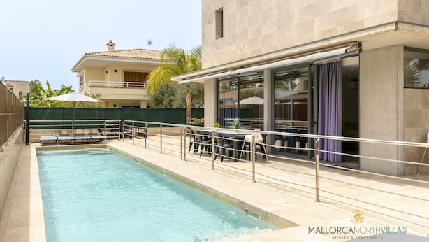 Una piscina con sillones de descanso y una mesa, un área de estar al aire libre techada, y un edificio con ventanas amplias y un balcón.