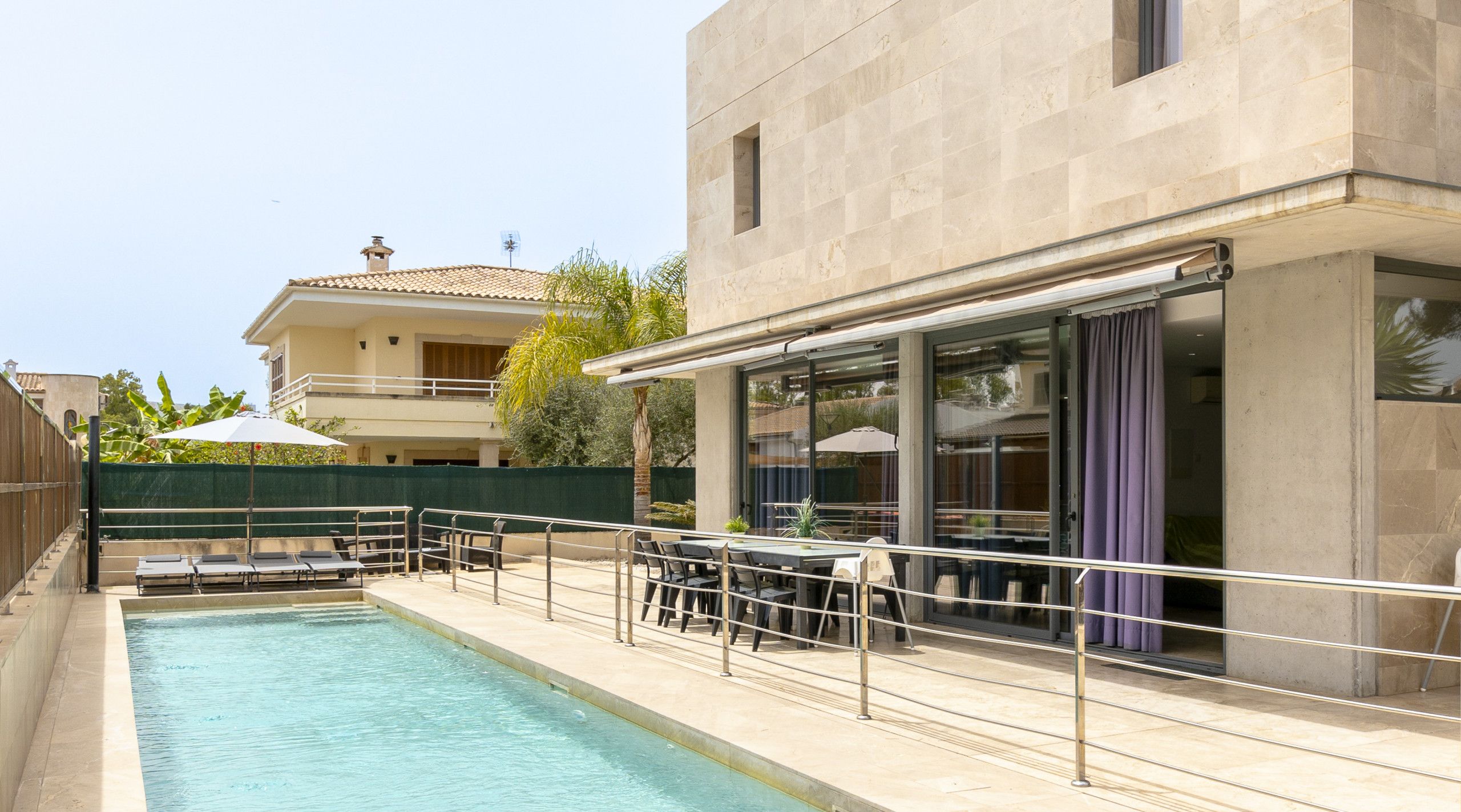 Una piscina con sillones de descanso y una mesa, un área de estar al aire libre techada, y un edificio con ventanas amplias y un balcón.