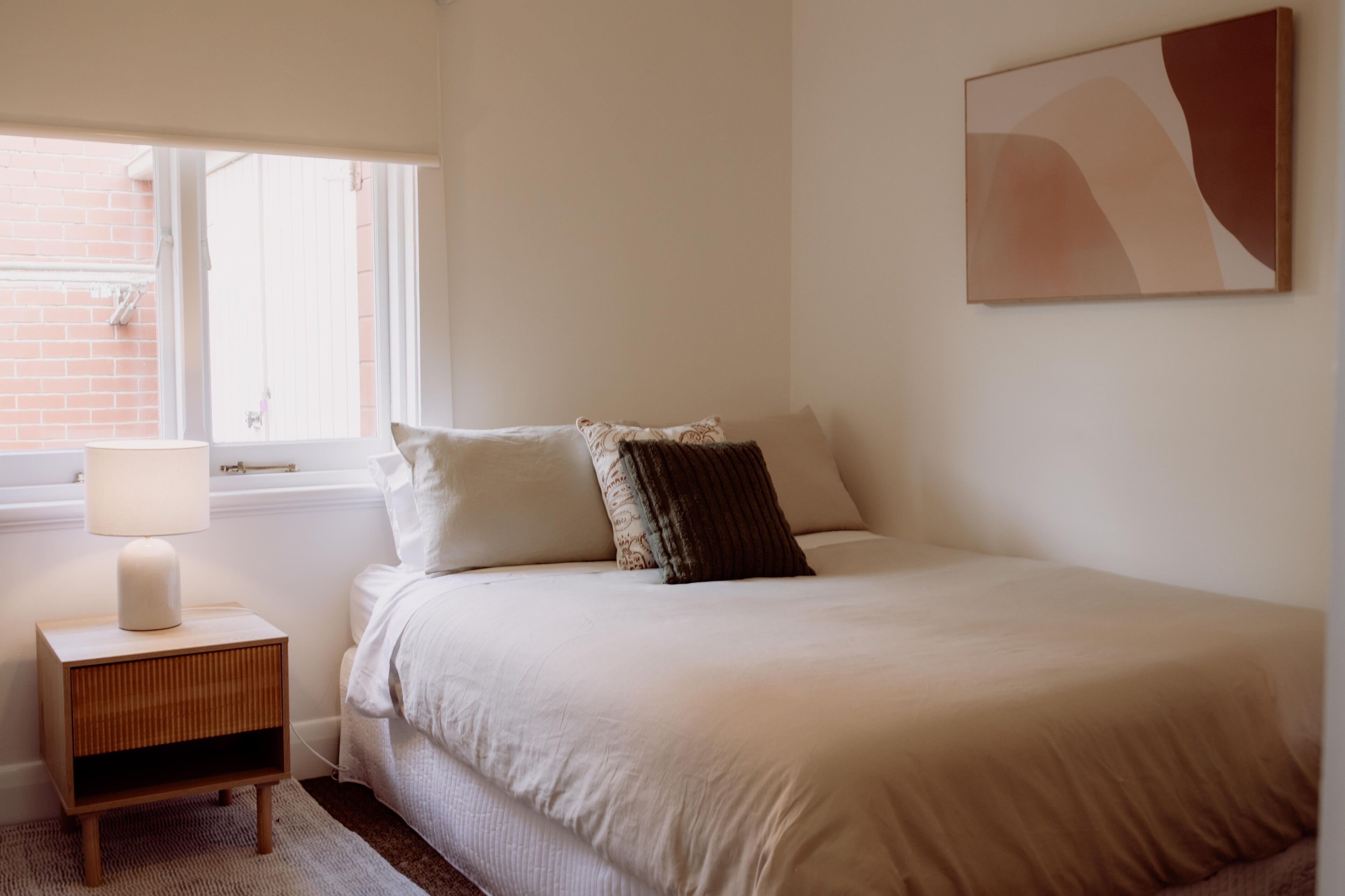 Een slaapkamer met een bed, nachtkastje met lamp, raam en een abstract schilderij aan de muur.