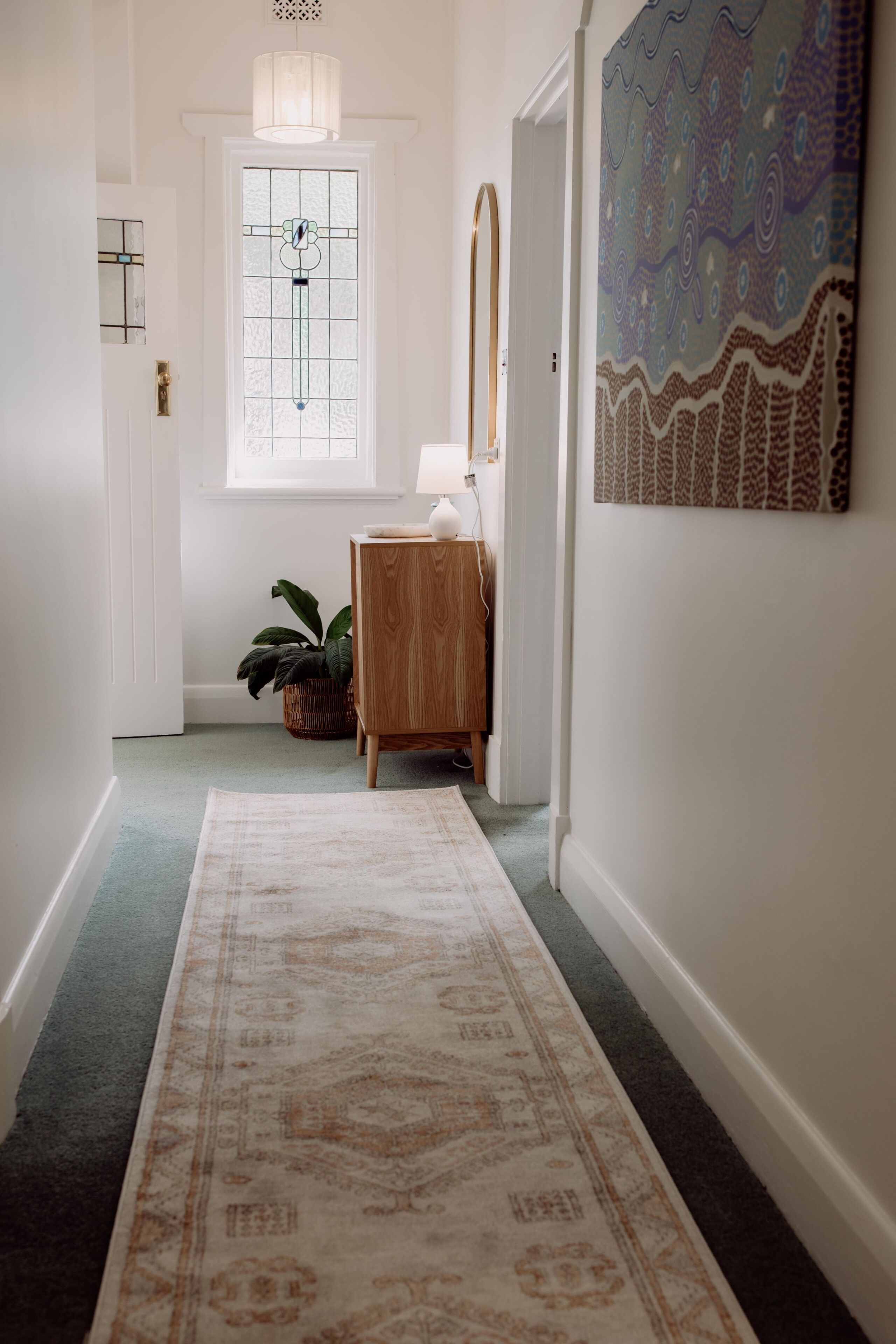 Un couloir avec un tapis à motifs, une commode en bois, une plante en pot, un miroir et un mur orné d’un tableau décoratif.