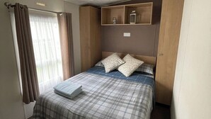 A compact hotel room with a bed, a small shelf with toiletries, a window with curtains, and a towel on the bed. - Caravan - Sleeps 10 - Pet friendly - Parking (Clacton-on-Sea)