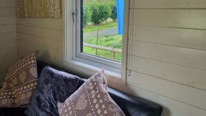 Een kamer met een bank tegen een muur, een raam met uitzicht op een tuin, en een geborduurde kussen op de bank.