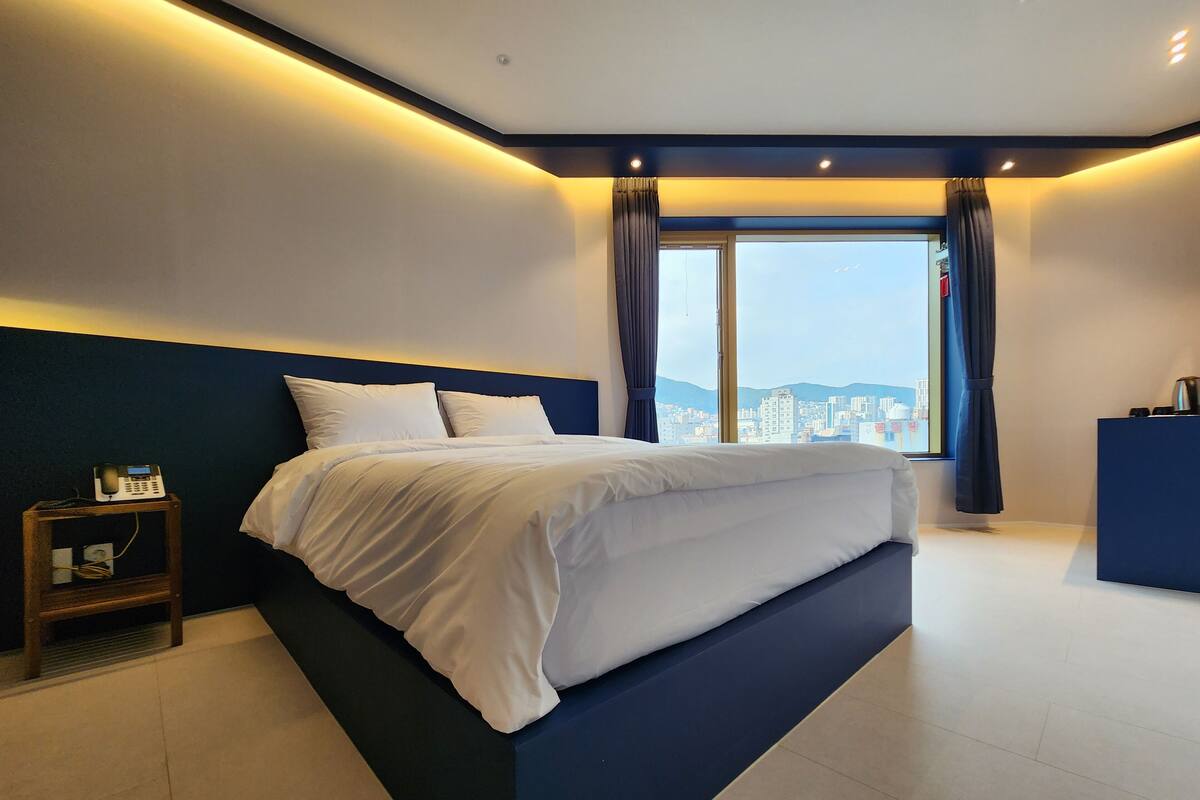 A modern hotel room with a large bed, a bedside telephone, and a view of the city through a large window.