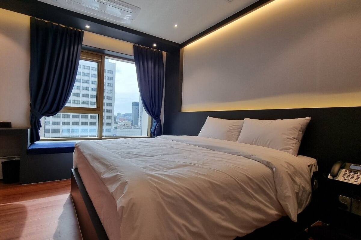 A modern hotel room with a large bed, a desk with a kettle, and a view of the city through a window with curtains.