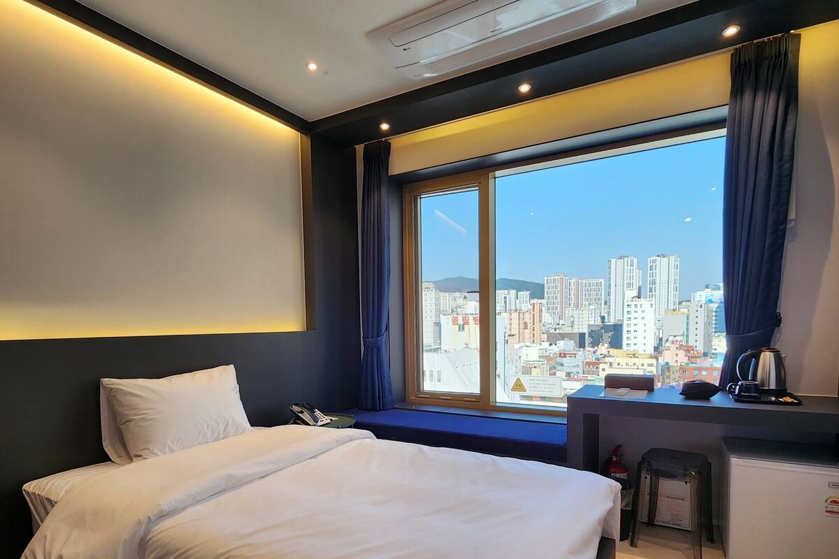 A hotel room with a bed, a desk, a window with a city view, and a coffee maker.