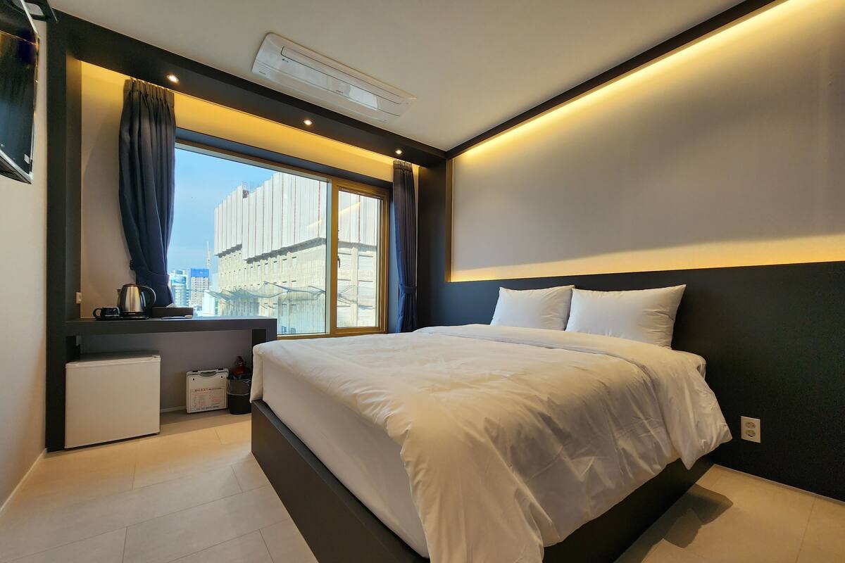 A modern hotel room with a large bed, a small desk with a kettle, a window with curtains, and a view of a cityscape.