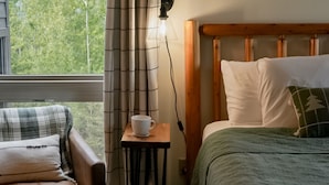 A bedroom with a wooden headboard, a bed with white bedding, a green throw blanket, a plaid chair with cushions, a small table with a cup, and a window with curtains. - Whirlpool Suite w/ Pool, Hot Tub, Hiking, Golfing, & More on Lutsen Mountain (Tofte)