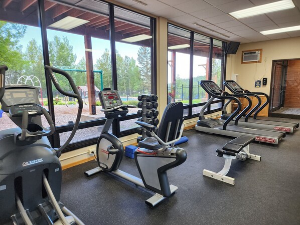 A gym with various exercise equipment, large windows, and a view of trees and a fence.