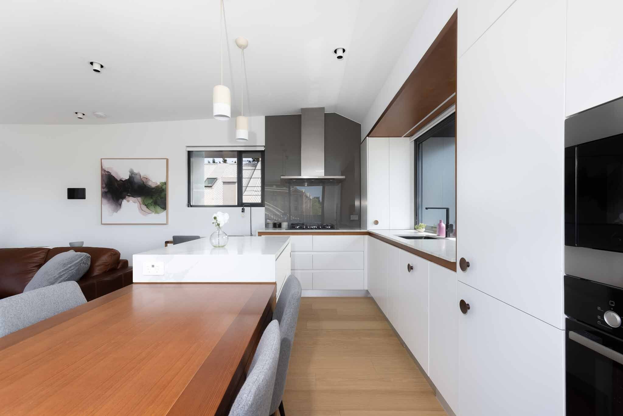 A modern kitchen with white cabinets, a wooden dining table, and a built-in oven.