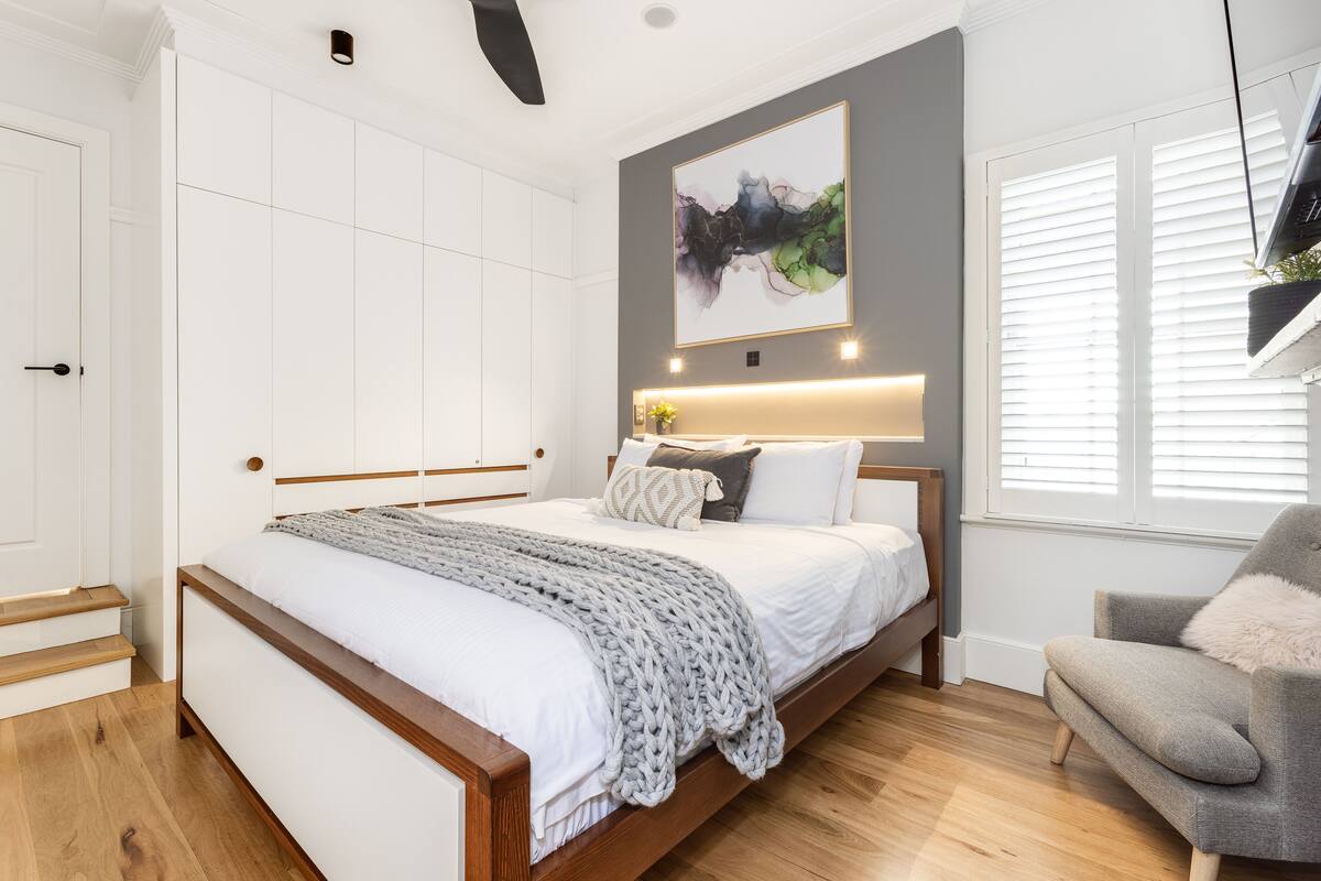 A bedroom with a wooden bed, white bedding, a grey chair, and a large abstract painting on the wall.