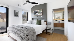 Une chambre moderne avec un grand lit, un ventilateur de plafond, un tableau encadré et une vue sur la ville par la fenêtre.