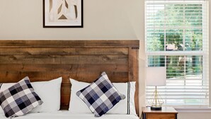 Una cama bien tendida con cabecera de madera, dos almohadas a cuadros, una mesita de noche con lámpara y una ventana con persianas.