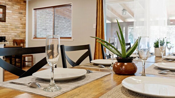 Un comedor con una mesa de madera dispuesta para cuatro, incluyendo platos, cubiertos y copas de vino. Hay una planta en maceta sobre la mesa, y un muro de ladrillo y una ventana al fondo.