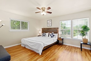 A bedroom with a wooden floor, a bed with white bedding, a ceiling fan, and two windows with blinds.