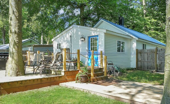 Situated in a lush green setting, this charming cottage features a wooden deck and a fenced area with mature trees and a well-maintained lawn.