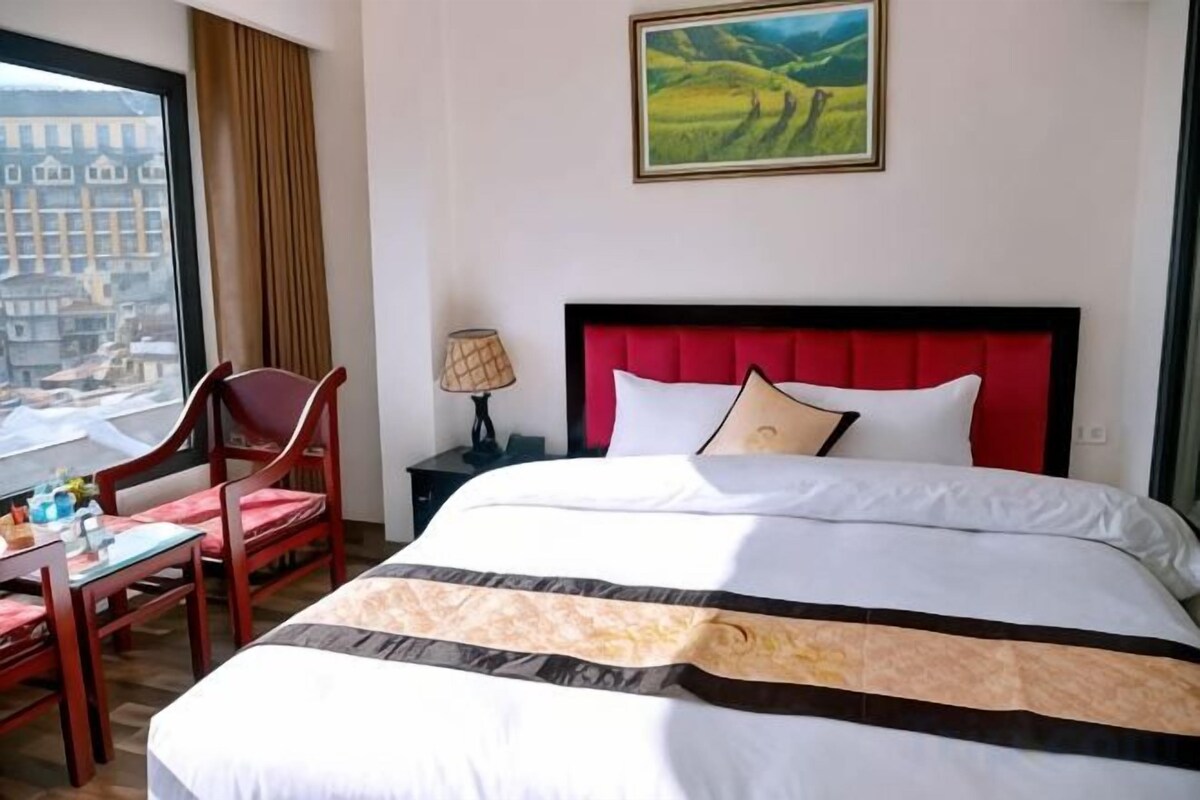 A hotel room with a large bed, a desk with a chair, a window with a view of buildings, and a painting on the wall.