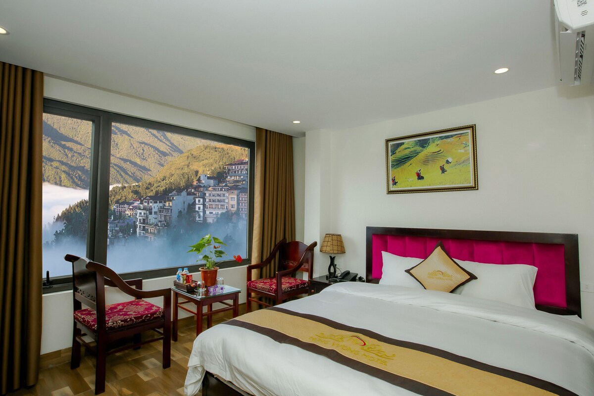 Habitación de hotel con una ventana grande, una cama, una silla, una mesa y un cuadro en la pared.