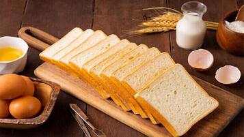 Slices of white bread on a wooden cutting board, surrounded by eggs, a bowl of flour, and a glass of milk.