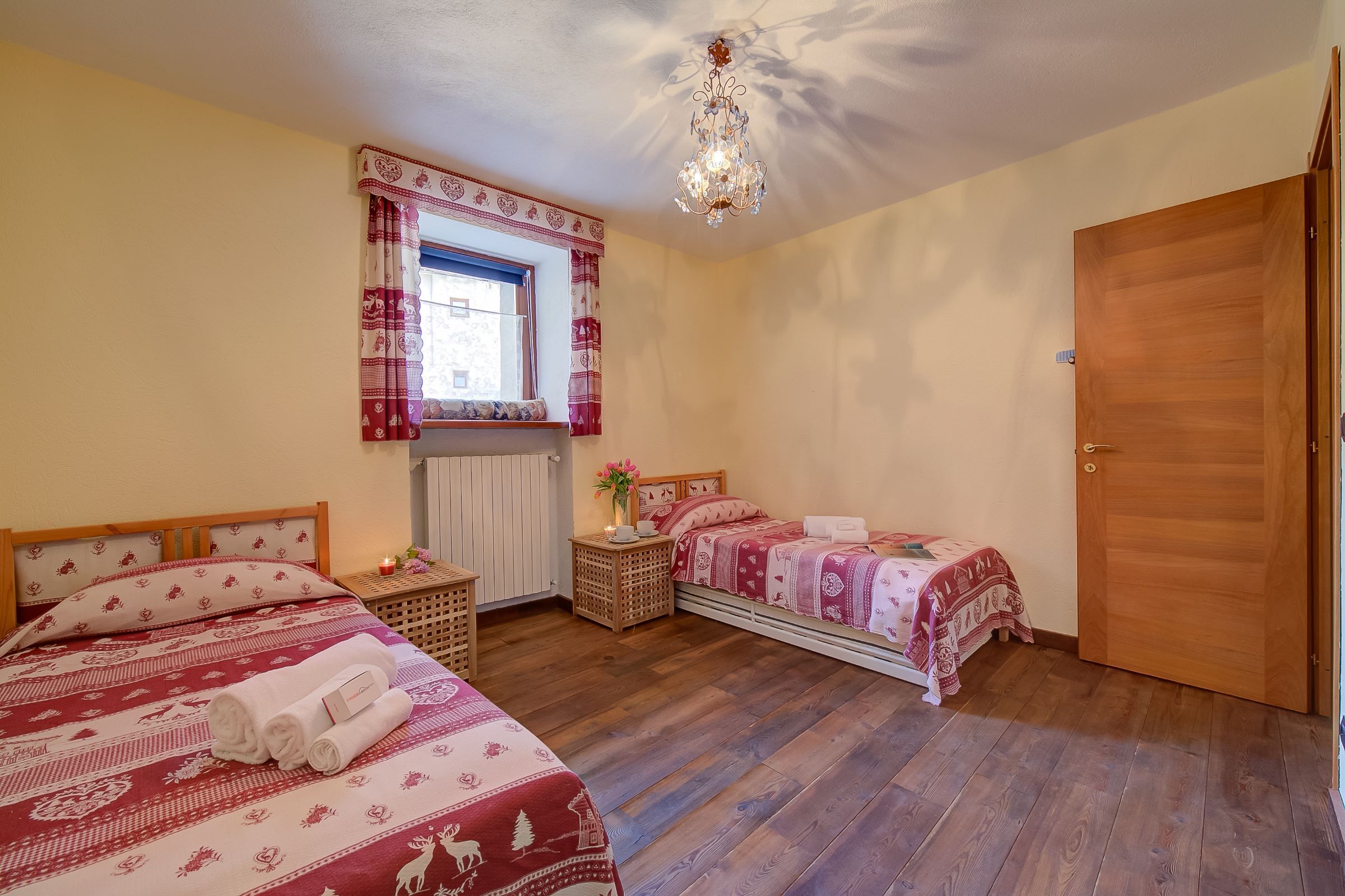 Un dormitorio con dos camas, suelo de madera, un candelabro, una ventana con cortinas y una puerta.
