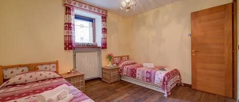 Un dormitorio con dos camas, suelo de madera, un candelabro, una ventana con cortinas y una puerta.