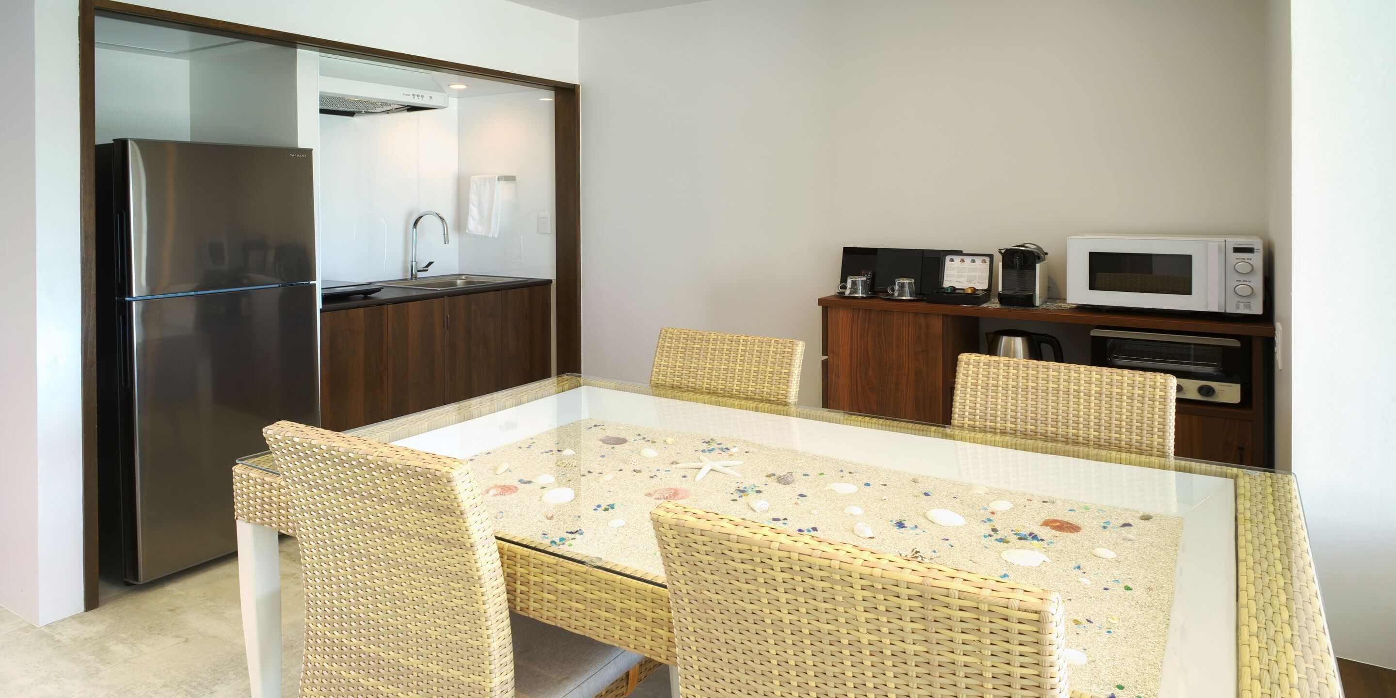 A compact kitchen with a refrigerator, microwave, and coffee maker. There is a glass-top table with wicker chairs.