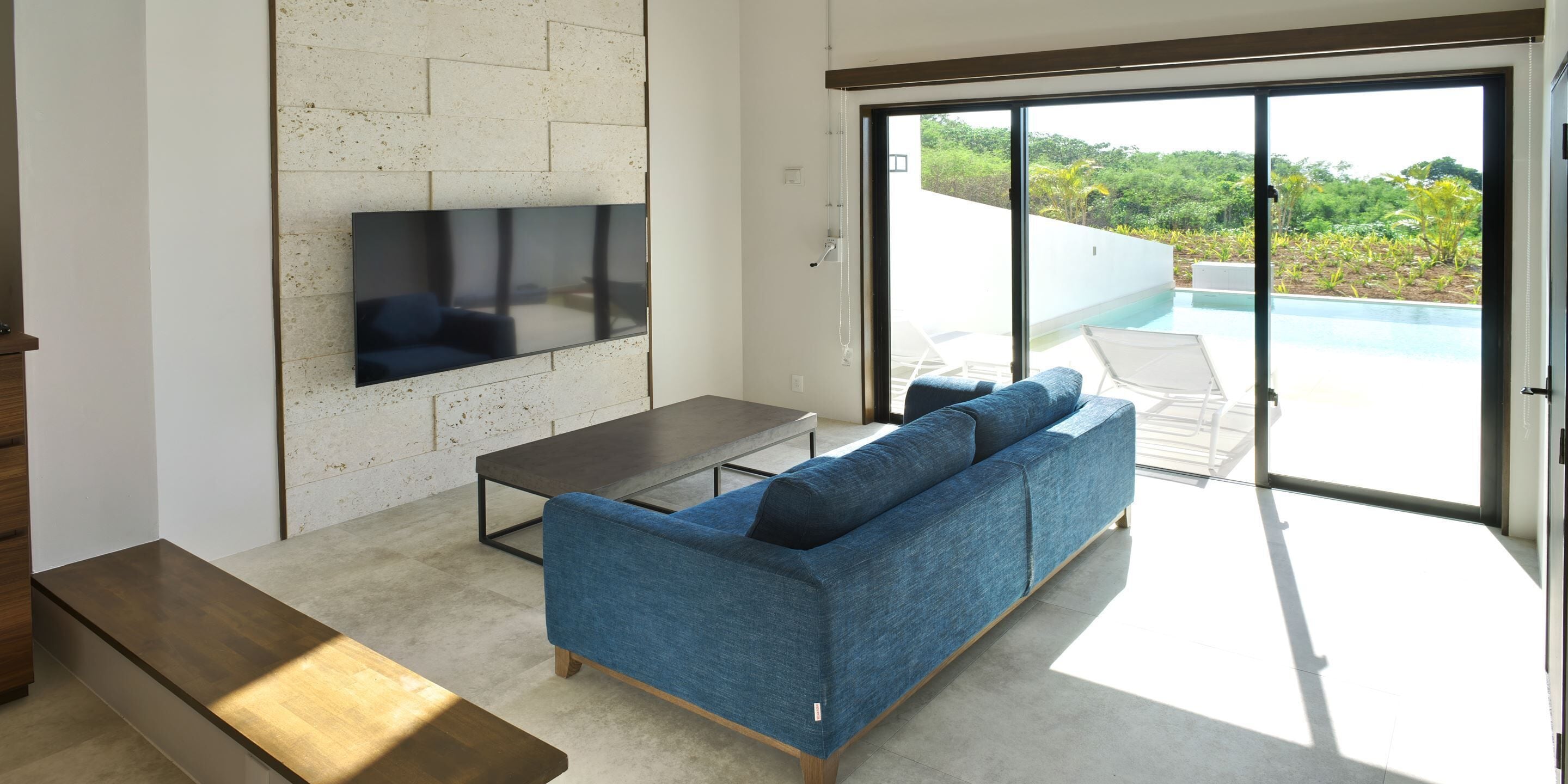 A modern living room with a blue sofa, a flat-screen TV mounted on a textured wall, a wooden bench, and a glass door leading to an outdoor pool area.