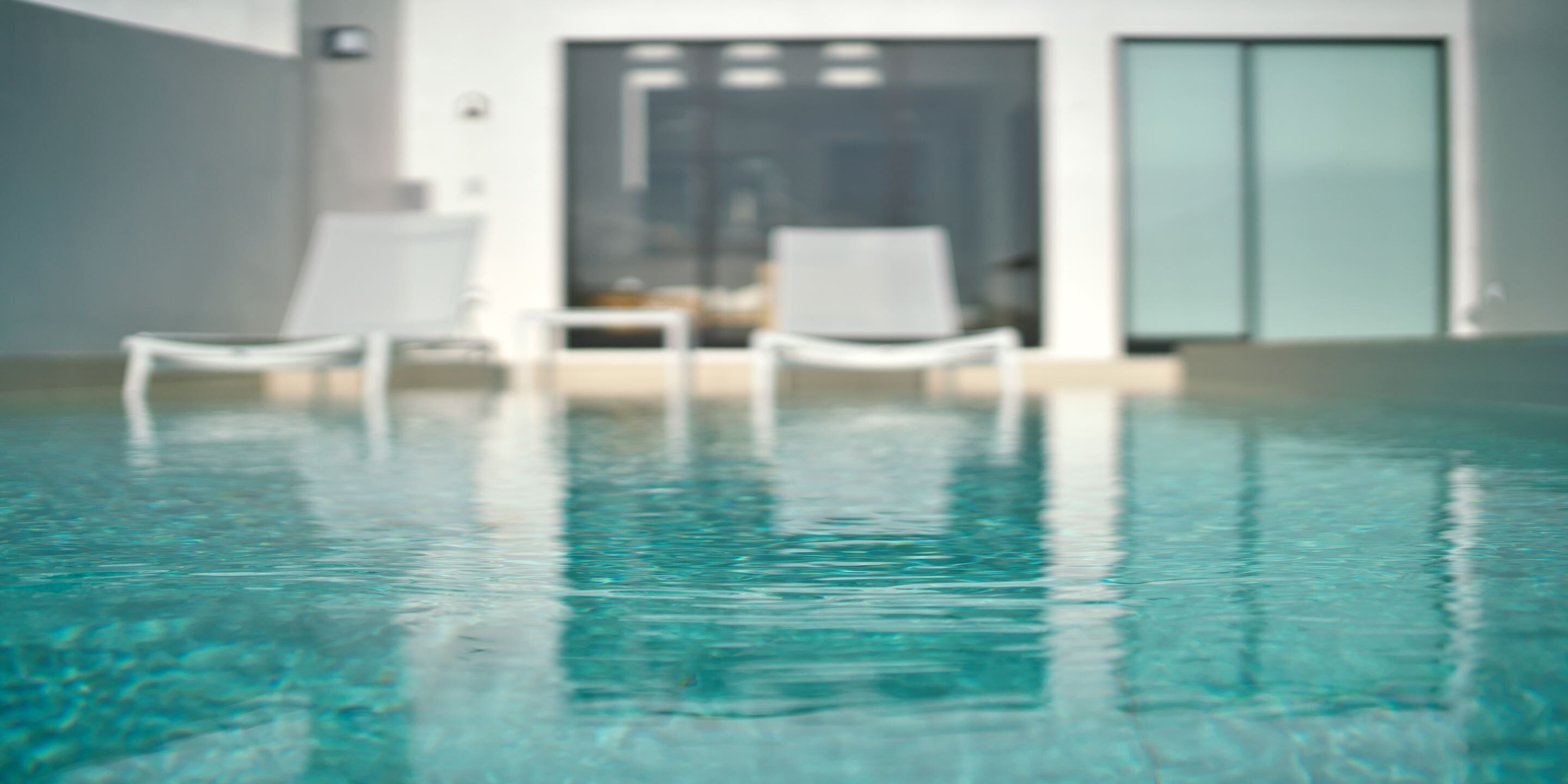 A clear indoor swimming pool with a modern design, featuring two white lounge chairs and a glass door leading to a room.