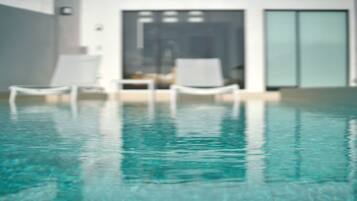 A clear indoor swimming pool with a modern design, featuring two white lounge chairs and a glass door leading to a room.