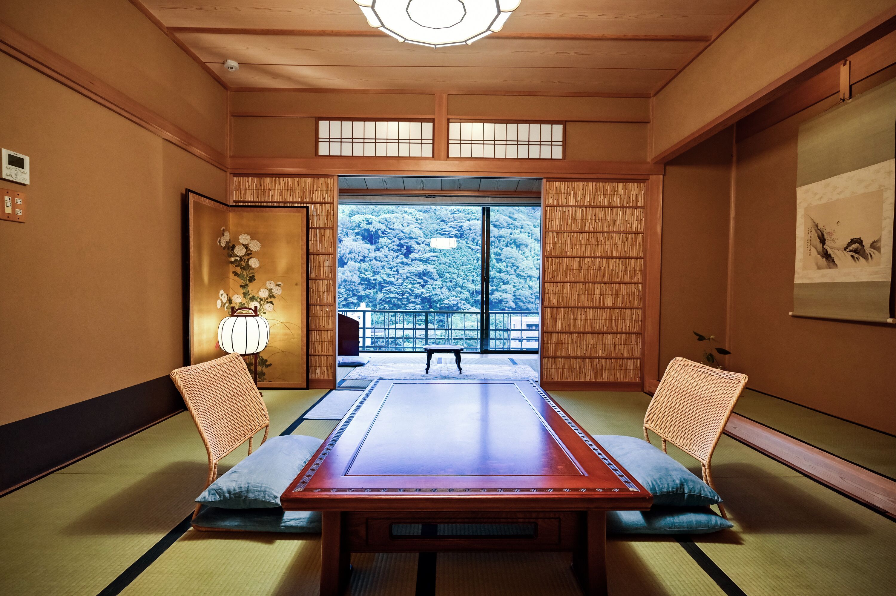 Ein traditioneller japanischer Raum mit Tatami-Boden, einem niedrigen Tisch, zwei gepolsterten Stühlen, einer hängenden Schriftrolle und Blick auf eine verschneite Landschaft durch große Fenster.