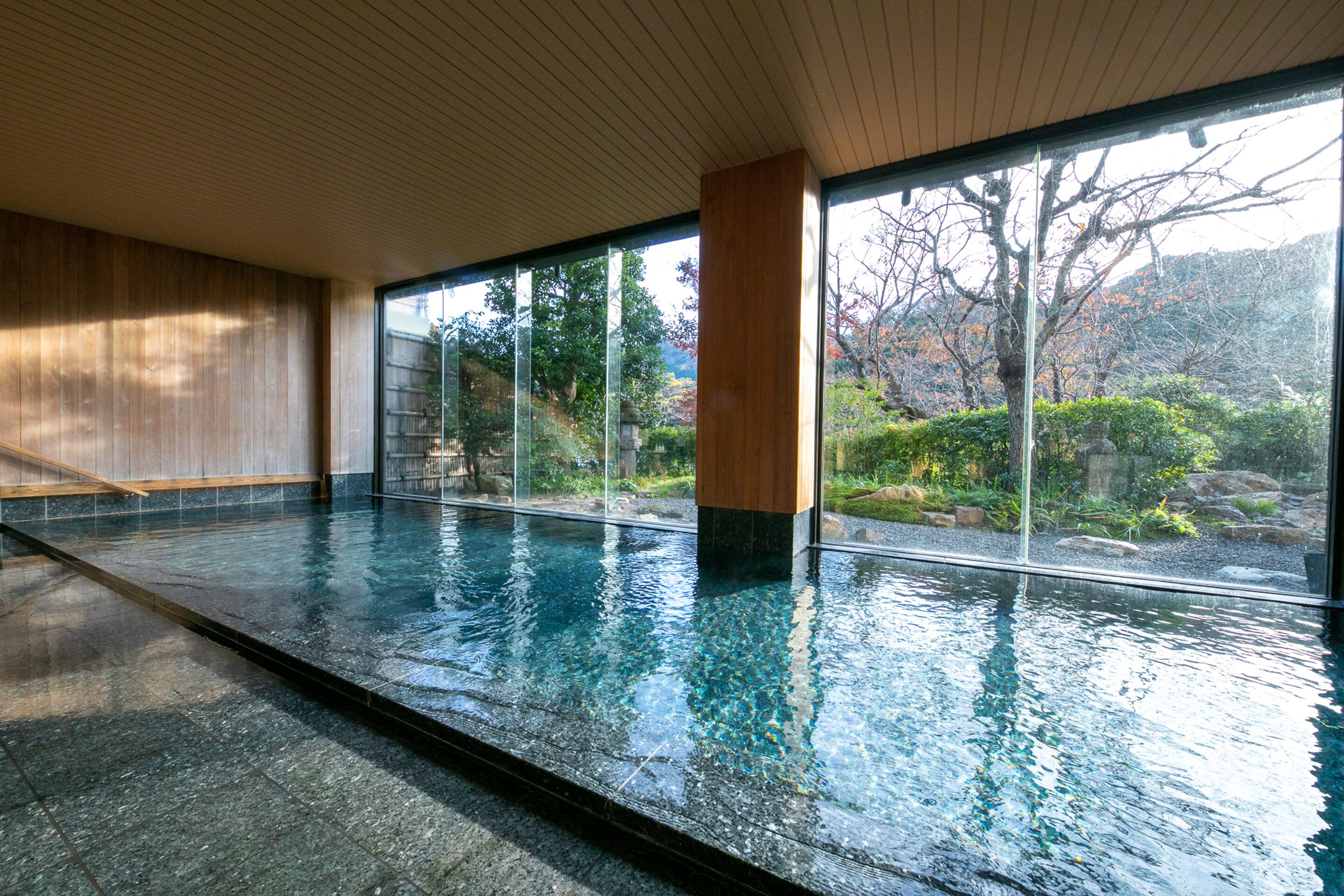 A large indoor swimming pool with clear glass walls, a wooden ceiling, and a view of a garden with trees and rocks.