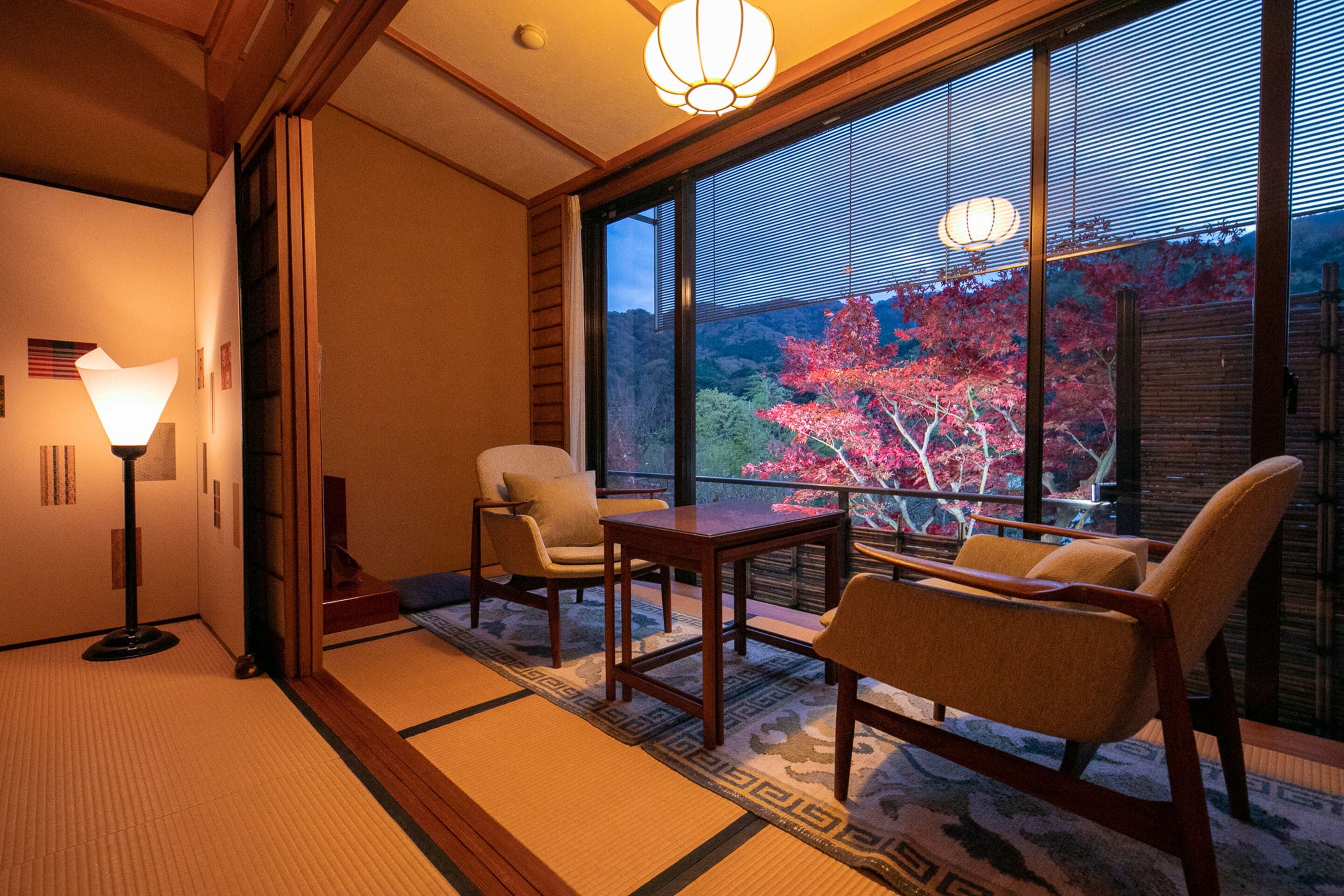 A traditional Japanese room with tatami flooring, a low table, two chairs, and a large window offering a view of autumn foliage.