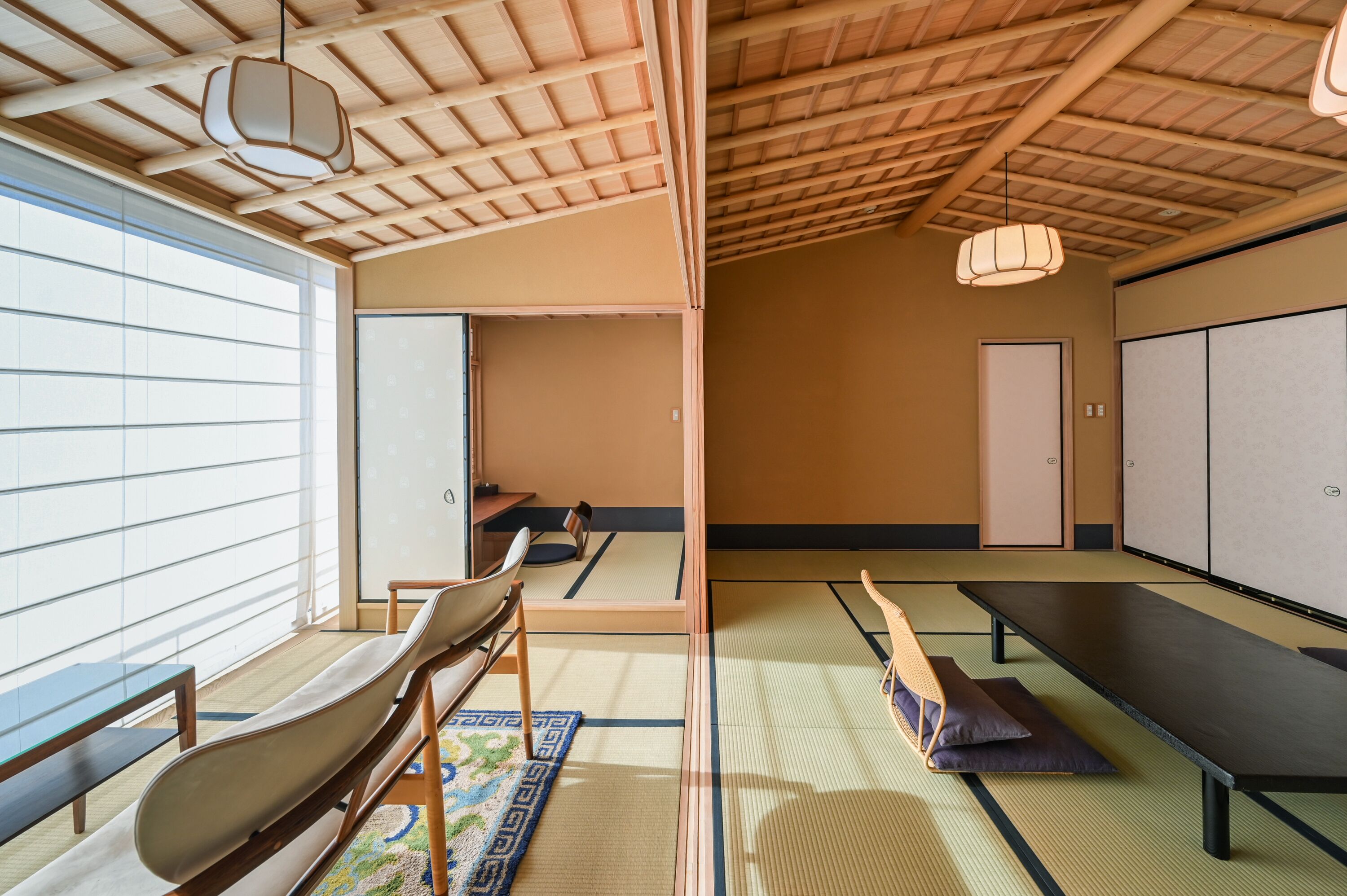 A tatami mat room with a low ceiling, wooden beams, tatami mats, a tatami mat table, a tatami mat bench, a hanging light fixture, and a sliding door.