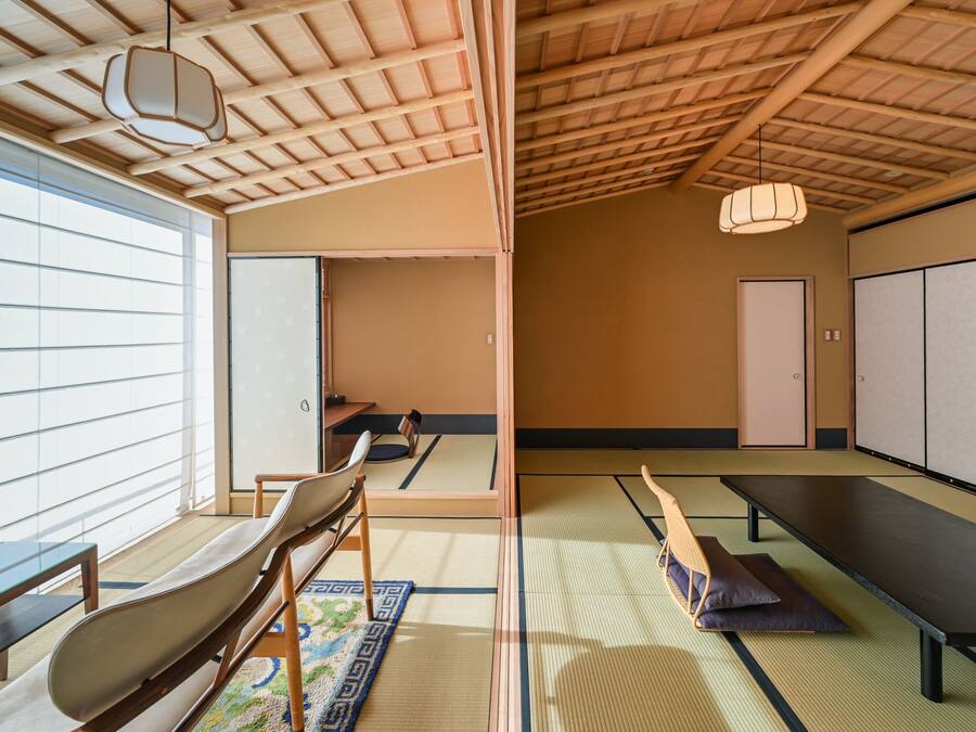 A tatami mat room with a low ceiling, wooden beams, tatami mats, a tatami mat table, a tatami mat bench, a hanging light fixture, and a sliding door.