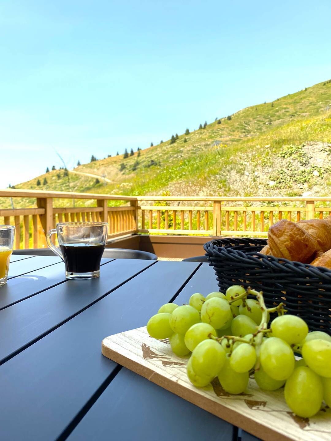 A table with a basket of green grapes, a glass of black coffee, and a croissant on a terrace with a wooden railing, overlooking a green hillside.