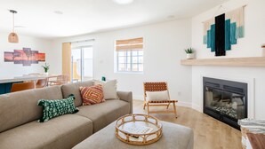Uma sala de estar moderna com um sofá, uma poltrona, uma lareira e uma zona de refeições ao fundo.