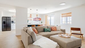 Uma sala de estar moderna com um sofá seccional, um banquinho pequeno e uma cadeira de madeira. Uma cozinha é visível ao fundo, com uma geladeira e uma área de jantar.