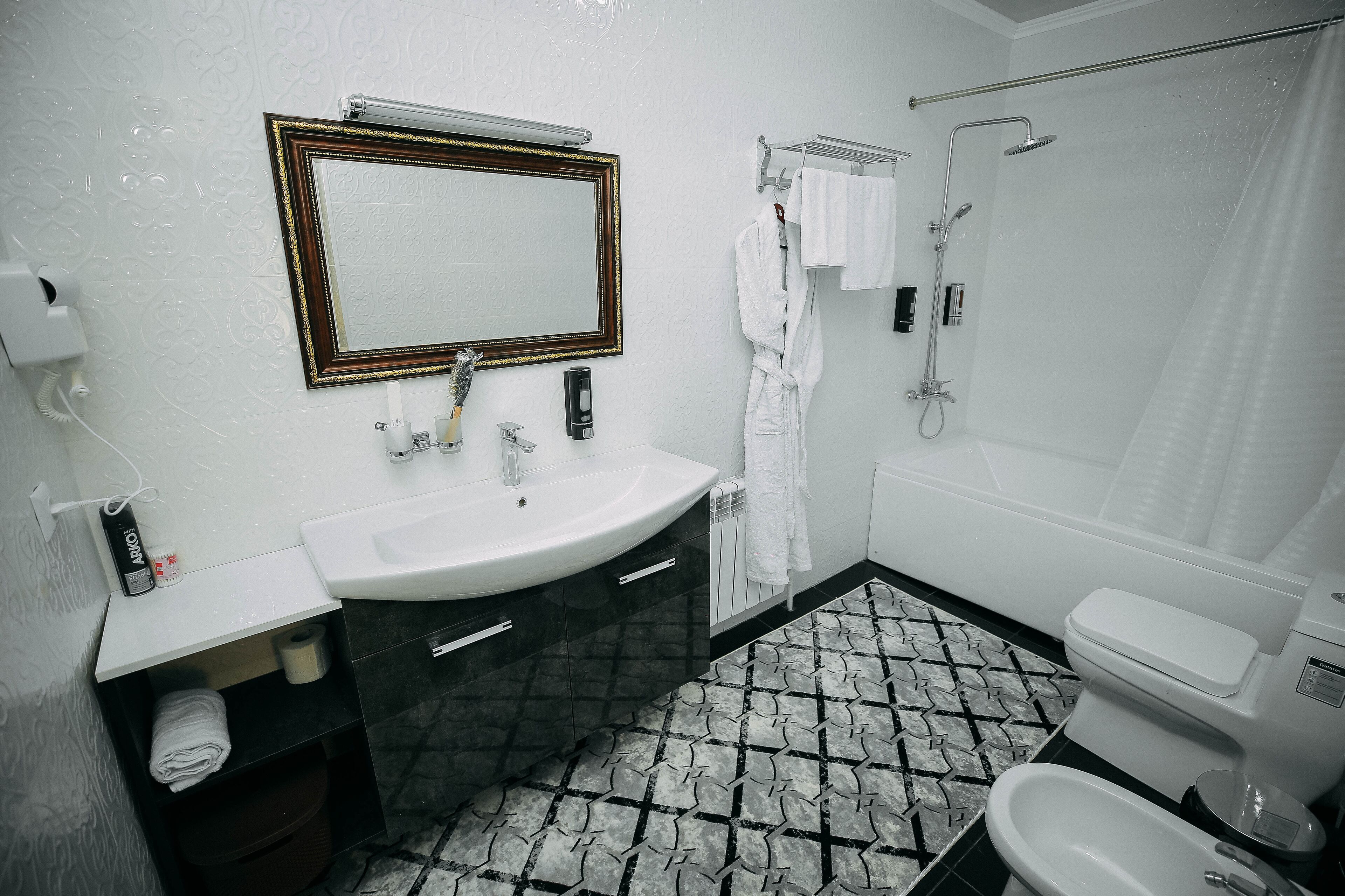 Un baño con lavamanos, espejo e inodoro blancos. Se ve un perchero con toallas, una ducha y un suelo estampado.