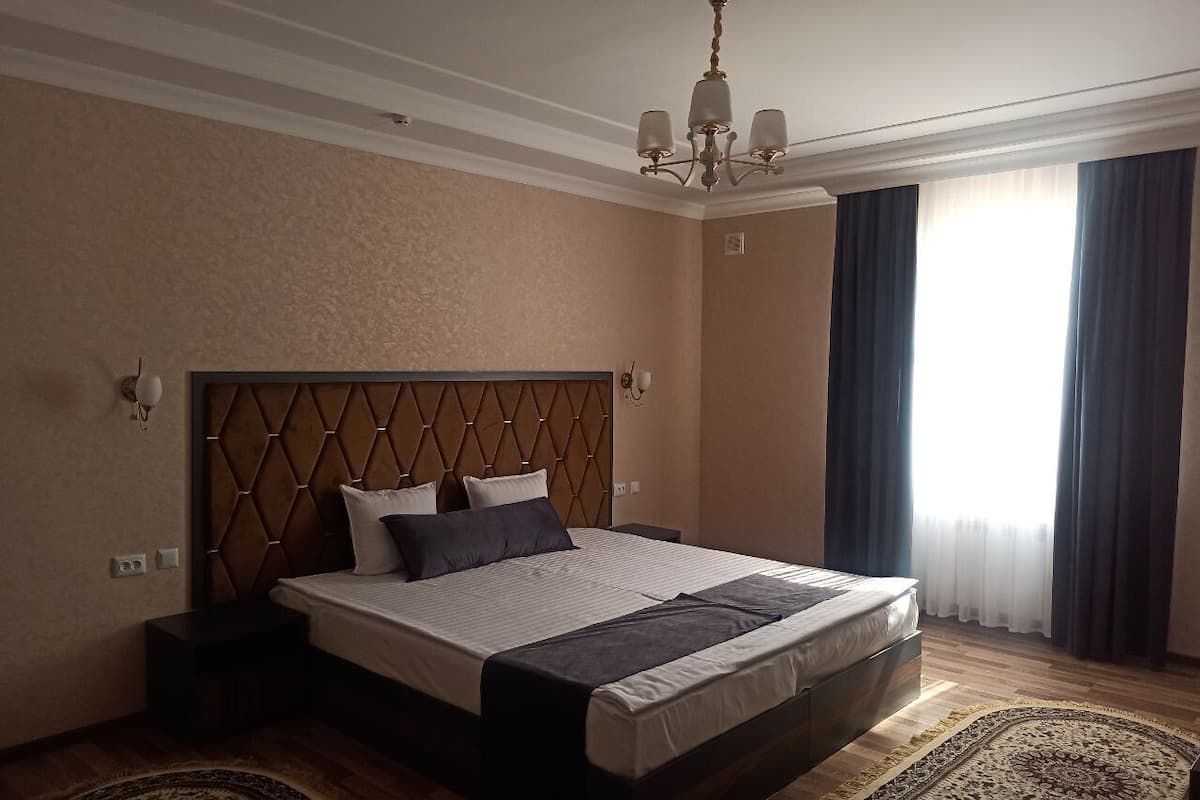 A hotel room with a large bed, a chandelier, a window with curtains, and a patterned rug.