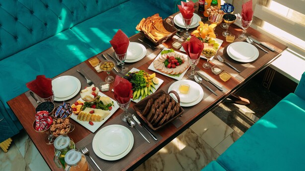 A breakfast table set with various dishes, drinks, and utensils in a bright room with a teal sofa. - FAYZ HOTEL (Urgench)