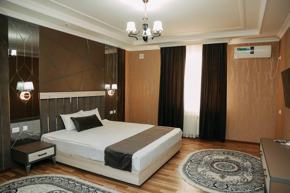 A modern hotel room with a large bed, bedside tables, a chandelier, an air conditioning unit, and a patterned rug.