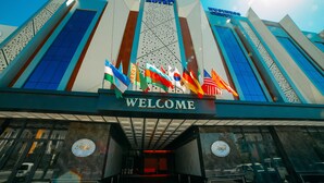 A hotel entrance with a 'WELCOME' sign, multiple flags, and a modern architectural design. - FAYZ HOTEL (Urgench)