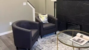 A living room with a dark armchair, a round glass coffee table, a patterned rug, a painting on the wall, and a flat-screen TV mounted above a fireplace. - Mr. Holmes Retreat (Pittsburgh)