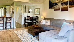 Ein modernes Wohnzimmer mit grauem Sofa, Esstisch aus Holz und Stühlen. Eine Treppe führt zu einer oberen Ebene.