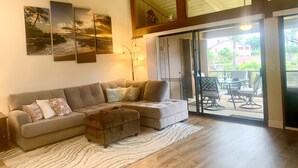 Ein Wohnzimmer mit Ecksofa, Couchtisch und Teppich. An den Wänden befinden sich Kunstwerke und eine Lampe. Das Zimmer hat eine Holzbalkendecke und eine Glasschiebetür, die zu einem Außenbereich mit Tisch und Stühlen führt.