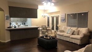 A living room with a dark wood floor, a white sofa, a black ottoman, and a dining area with a table and chairs.