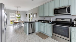 A modern kitchen with light blue cabinets, stainless steel appliances, and a dining area with white chairs. - Wonderful Ocean Views - Sleeps 12 -3 Mins to Beach (Oak Island)
